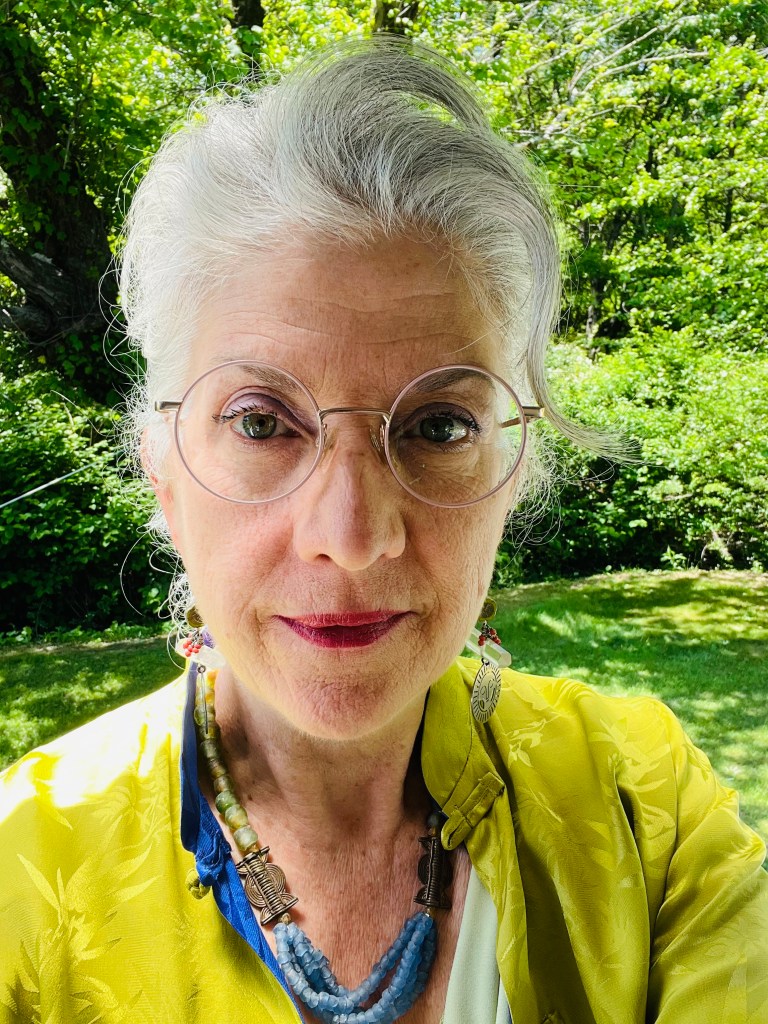 A close-up portrait of a woman with silver hair, wearing glasses and colorful jewelry, standing outdoors in a lush green setting.